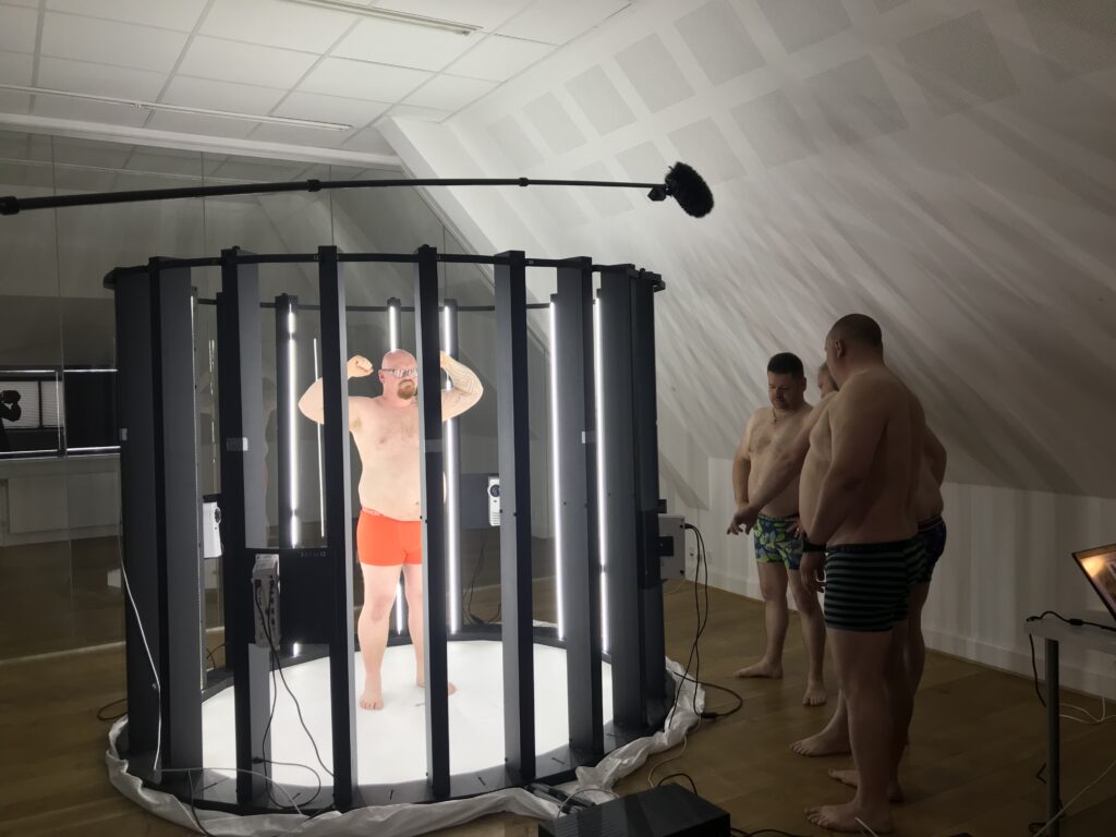Scanning af person til print af 3D figur. Der står tre personer udenfor kamerarummet. De fire personer deltager i TV programmet "rigtige mænd".